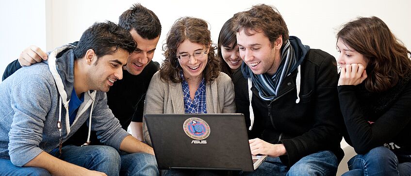 Sechs Studierende diskutieren über einen am Laptop gezeigten Inhalt.