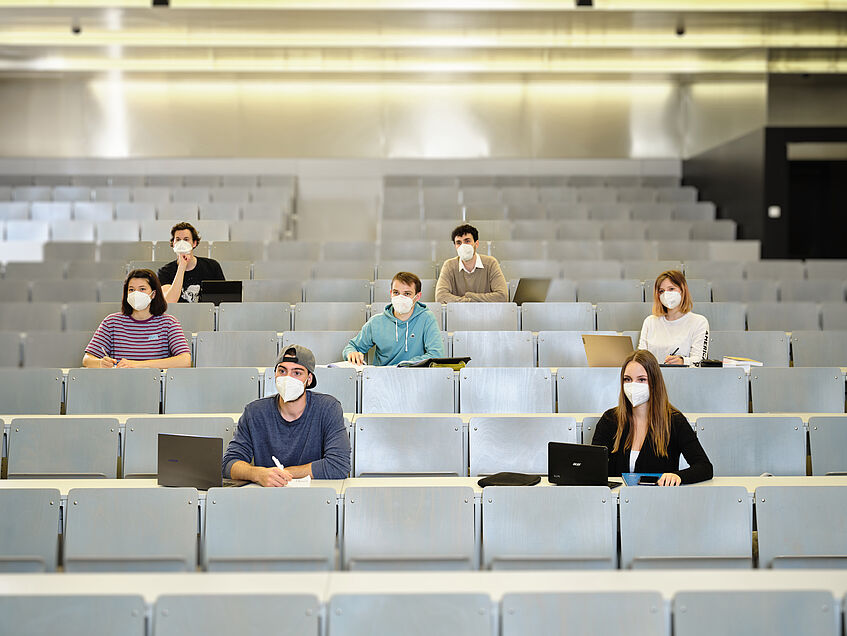 Studierende sitzen im Schachbrettmuster und mit FFP2-Maske im Hörsaal.