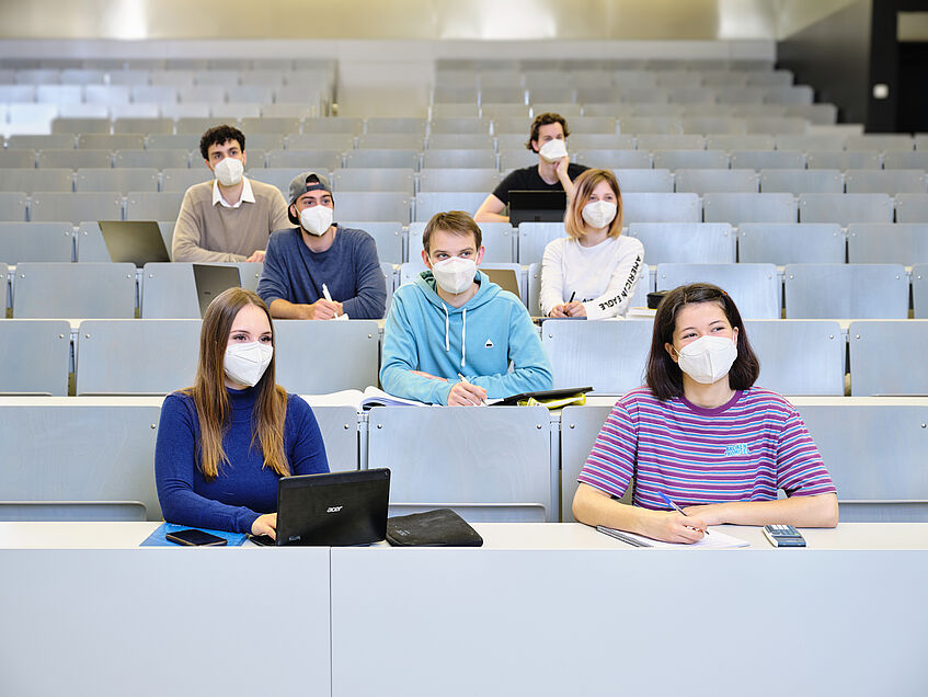 Studierende sitzen im Schachbrettmuster mit FFP2-Maske im Hörsaal.