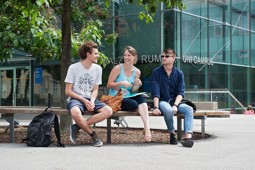Three students sitting on a bench in front of the Hörsaalzentrum at the Campus of the University of Vienna