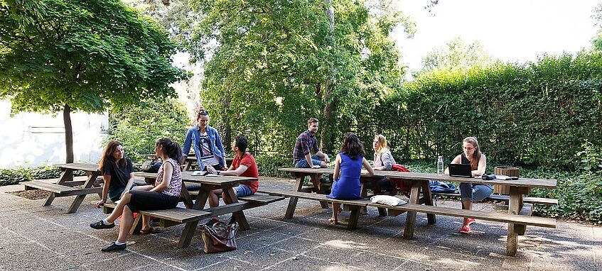 Studierende sitzen im Freien auf bzw. an Tischen.