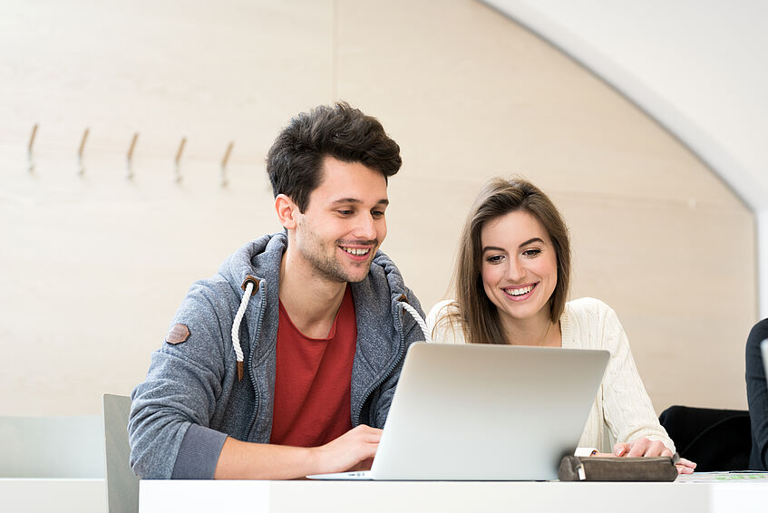Student und Studentin sitzen gemeinsam an einem Laptop und blicken auf den Bildschirm.