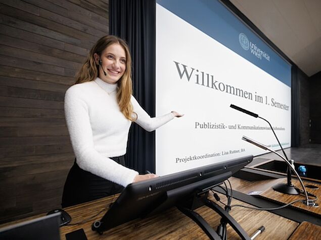 Junge Frau an einem Rednerpult zeigt auf eine Präsentation.