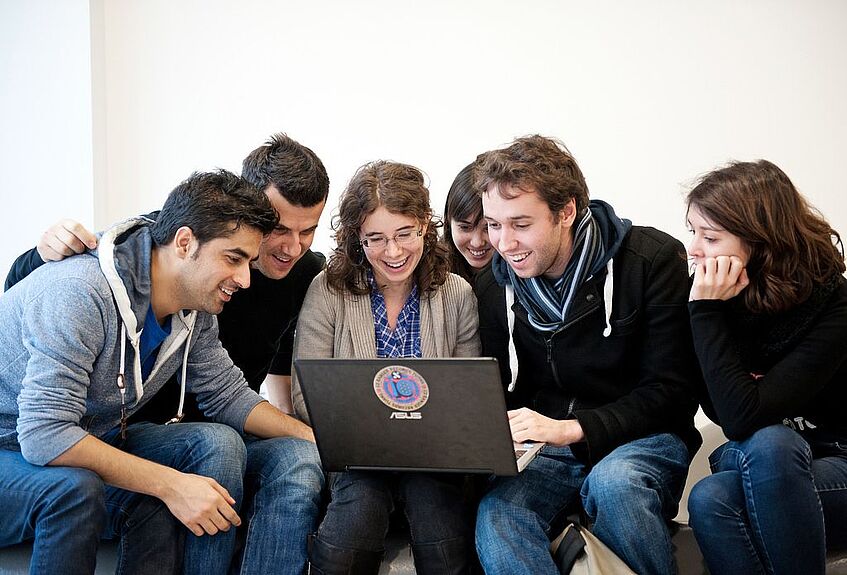 Mehrere Studierende und eine Lehrerin sitzen um einen Laptop herum.