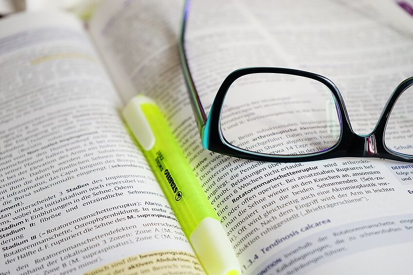 Eine Brille liegt auf einem aufgeschlagenen Buch, welches mit einem Leuchtstift offen gehalten wird.